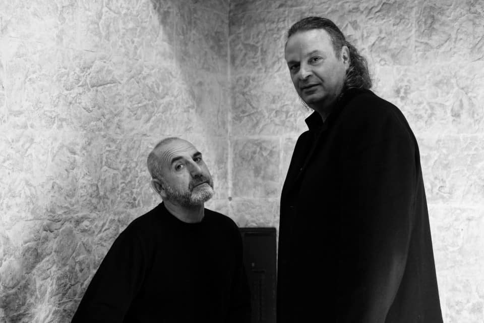 Άνδρες σε μουσική εκδήλωση, ένας με γκρίζα γενειάδα και ο άλλος με μακριά μαλλιά, στέκονται κοντά σε.